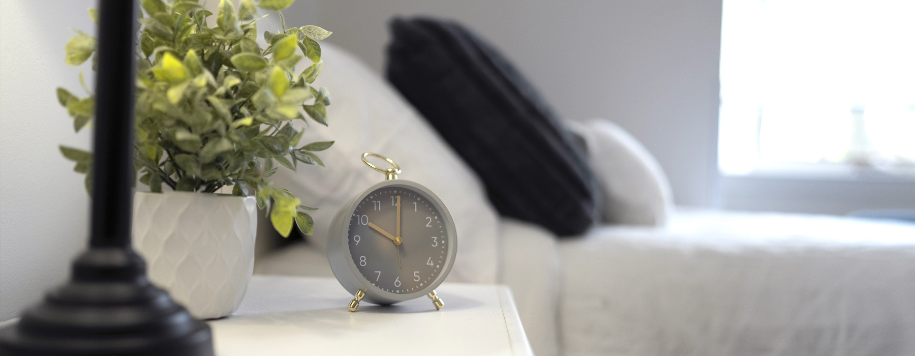 Closeup across night stand with lamp and alarm clock beside bed covered in white linens